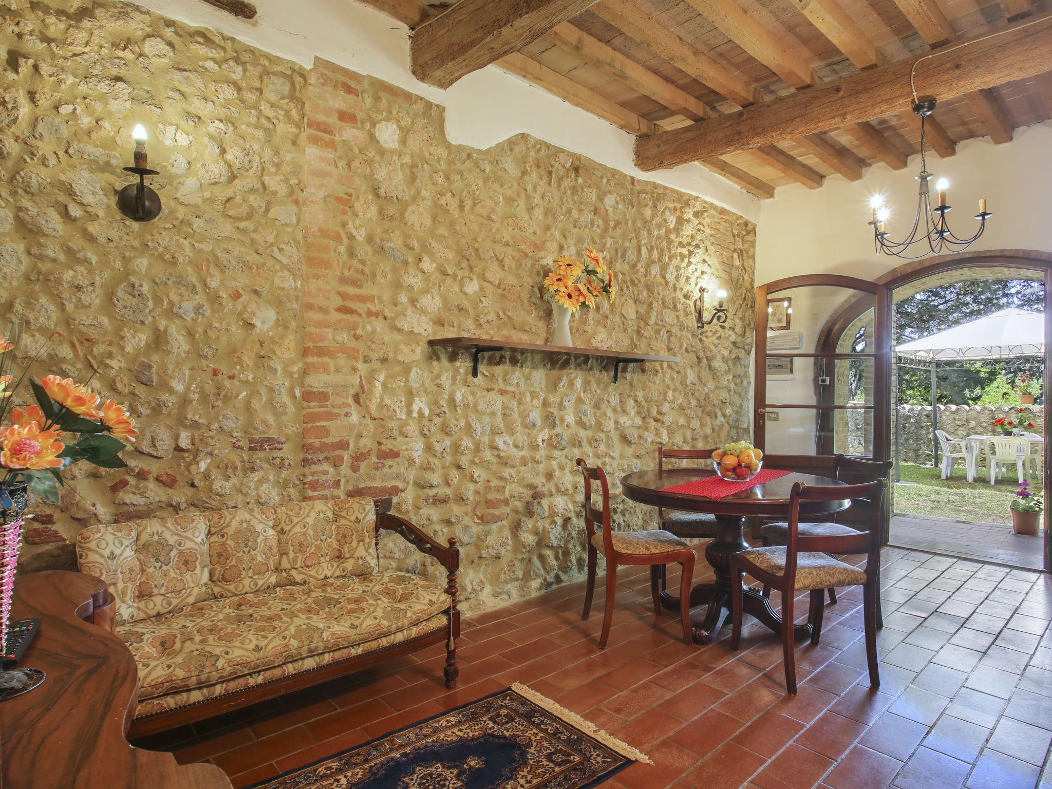 Una habitación acogedora con pared de piedra, vigas de madera en el techo, una mesa redonda para comer con sillas, un sofá y un jarrón con flores.