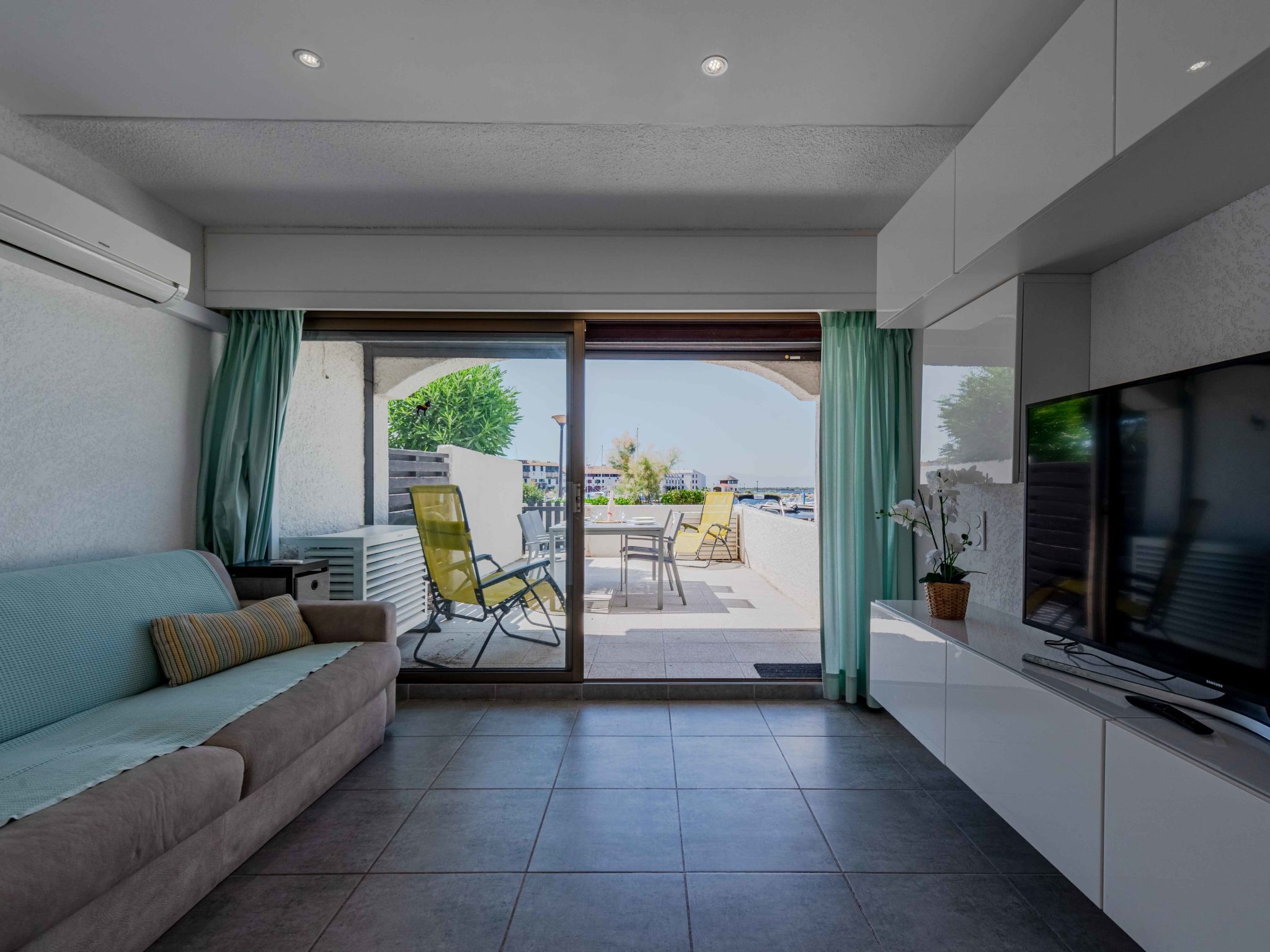 Ein modernes Wohnzimmer mit einer Couch, einem Fernseher und einer Schiebetür zu einer Terrasse im Freien mit Stühlen und Meerblick.
