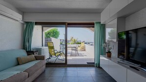 Een moderne woonkamer met een bank, een televisie en een schuifpui naar een terras buiten met stoelen en uitzicht op zee.