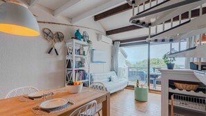 A cozy living room with a wooden dining table, white chairs, a bookshelf, and a large window offering a view of the sea. - Antigua by Interhome (Le Barcarès)