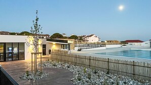 A modern outdoor pool area with a wooden deck, a small garden, and a building with a glass facade. - Antigua by Interhome (Le Barcarès)
