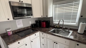 Una cocina con armarios blancos, microondas, lavabo y ventana con persianas.