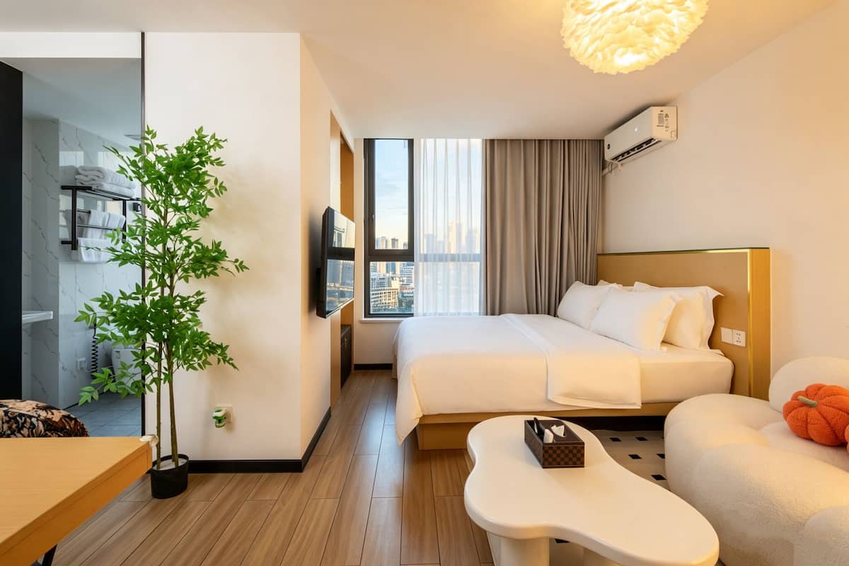 A hotel room with a bed, a TV, a plant, a window with a city view, and a desk.