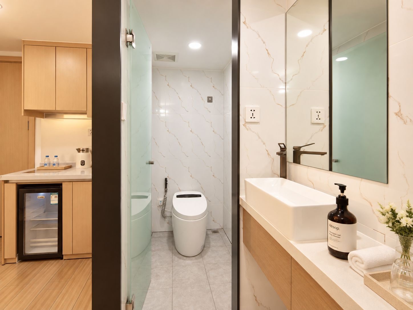 A modern bathroom with a glass shower enclosure, a white toilet, a sink with a large mirror, and a bottle of Aesop body wash.