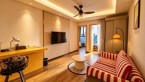 A modern living room with a sofa, a TV, a ceiling fan, and a view of the city. - Amanda Hotel (Phnom Penh)