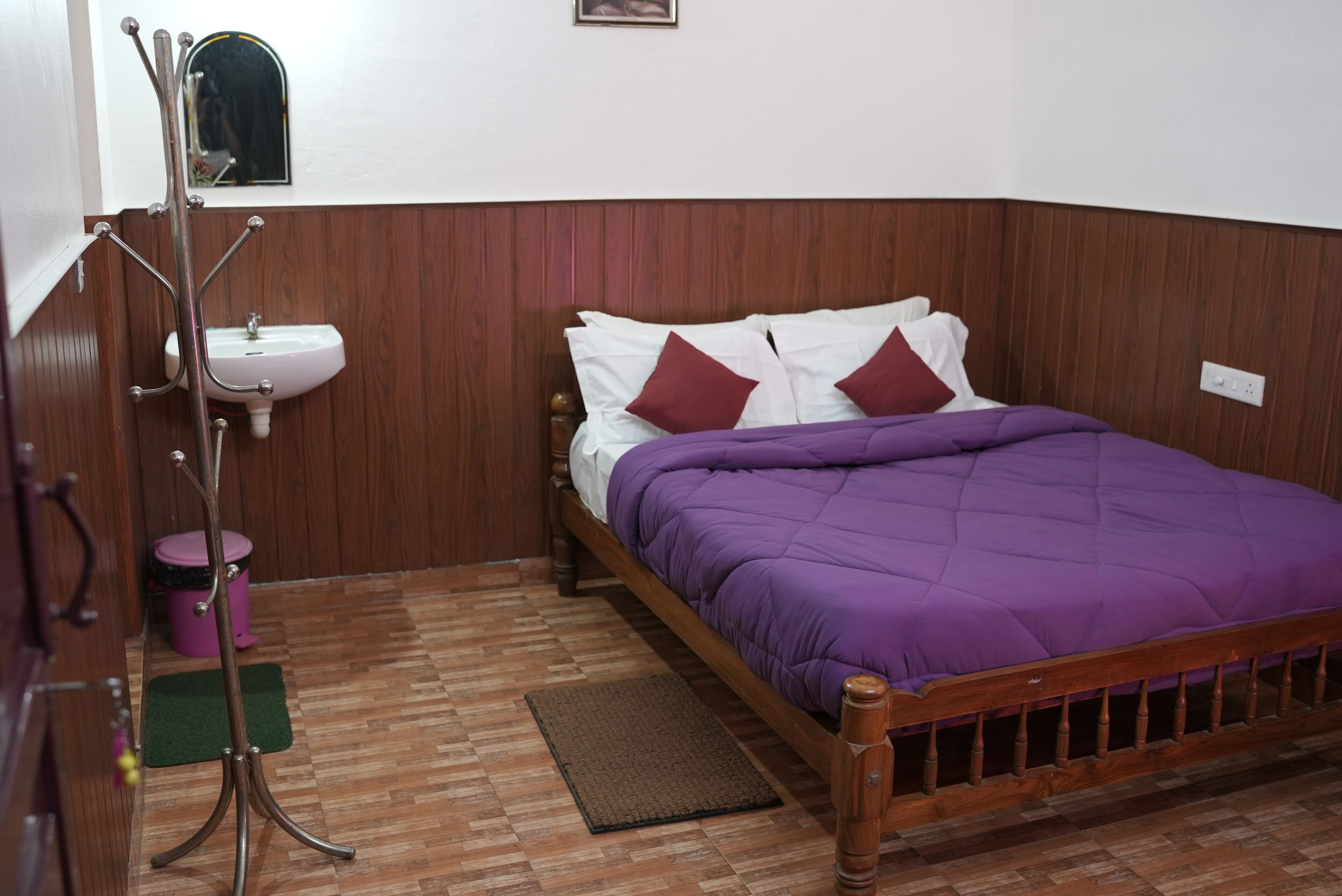 Een slaapkamer met een houten bed, paars beddengoed, een houten hoofdbord, een witte muur, een houten vloer, een nachtkastje van hout, een spiegel, een wastafel en een groene mat.