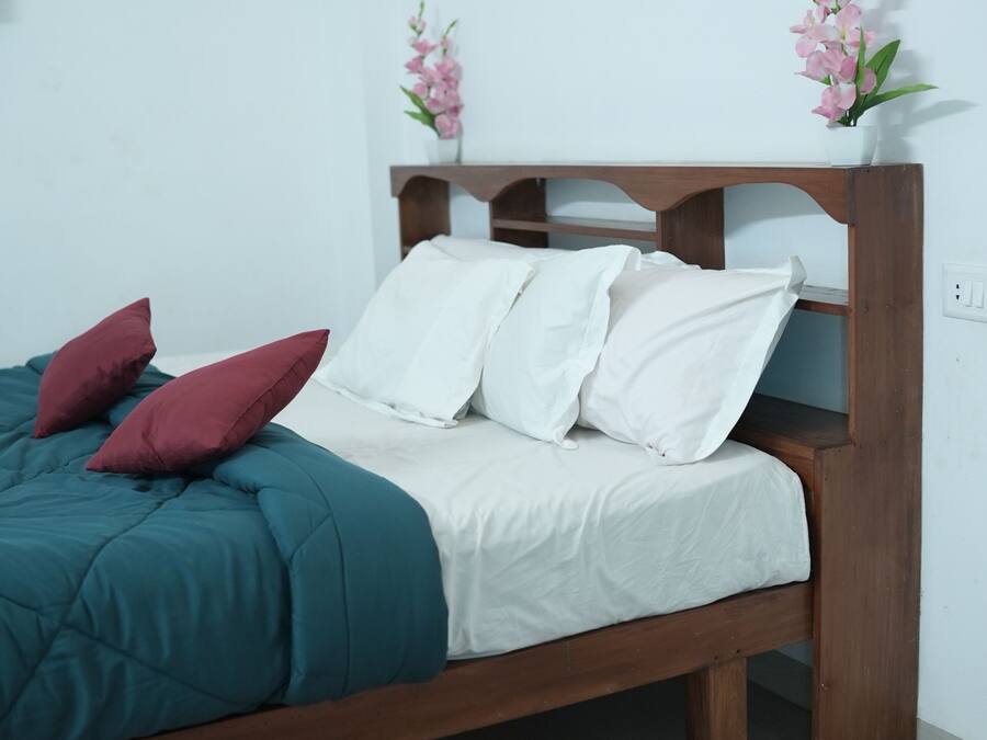 Una cama con edredón verde azulado, sábanas blancas y almohadas color burdeos. Tiene un cabecero de madera, un tapete a rayas rojas y rosas, y flores rosadas en un jarrón sobre la mesita de noche.