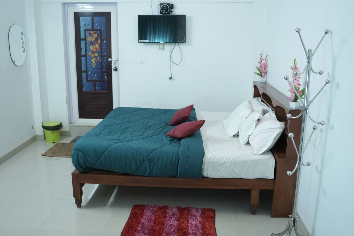 Ein Schlafzimmer mit einem hölzernen Bett, türkis-weiß gestreifter Bettwäsche, einem roten Teppich, einer grünen Mülltonne und einem an der Wand montierten Fernseher.
