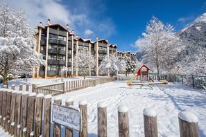 A snow-covered outdoor area with a wooden fence, a playground, and a building with multiple balconies. - Grand Roc A348 150m from Slopes & Parking, Argentière (Chamonix), France (Argentière (Chamonix))