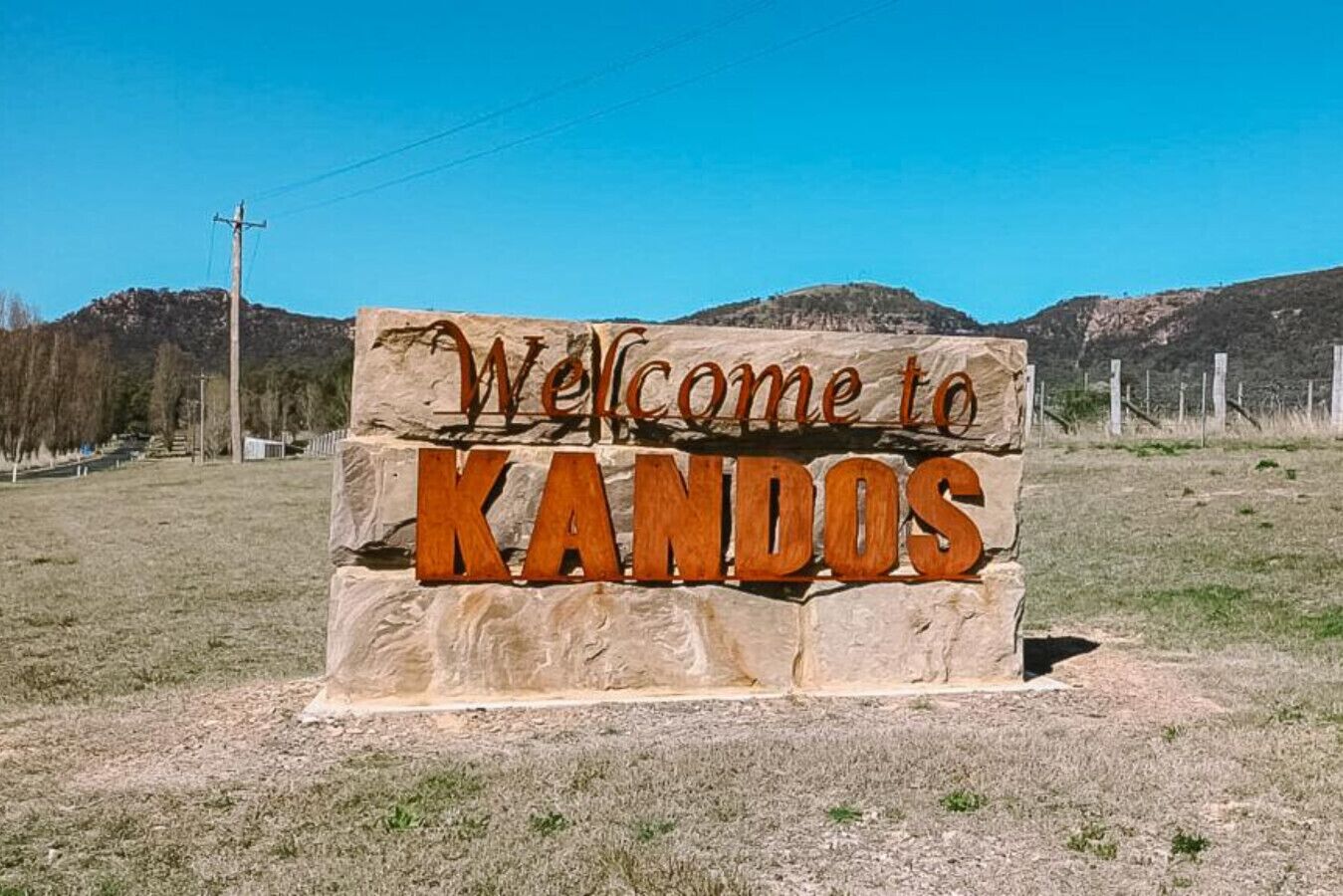 Señal de bienvenida a KANDOS con cielo despejado y montañas al fondo.