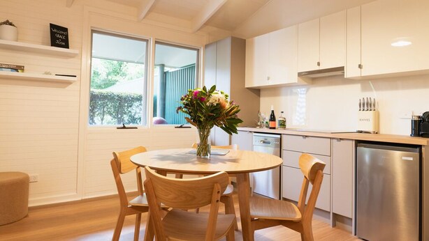 Una cocina moderna con armarios blancos, una mesa de comedor con sillas, un florero con flores y vistas al exterior a través de una ventana.