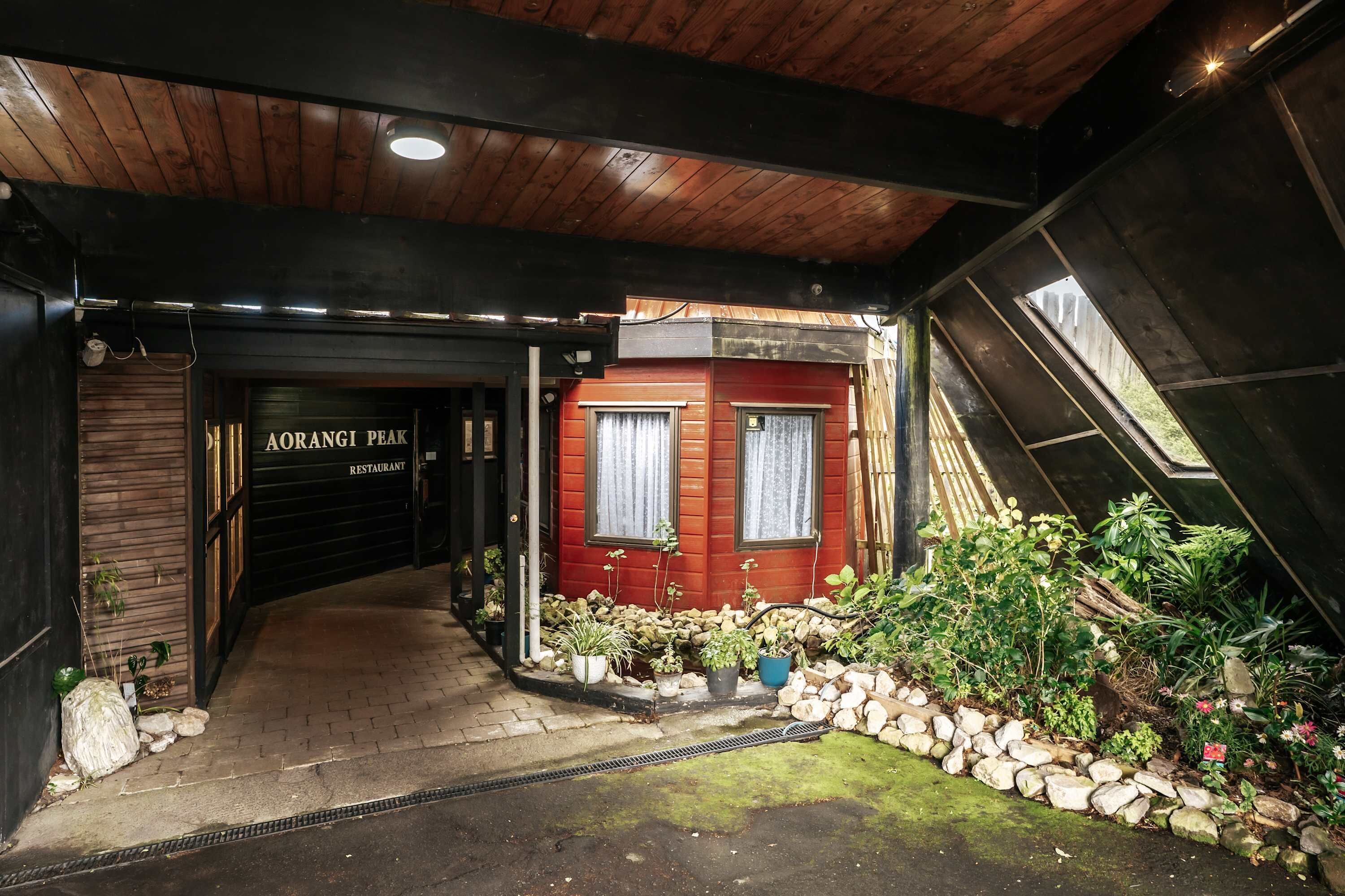 Entrada de un restaurante con techo de madera, un edificio rojo y un área ajardinada con plantas y rocas.