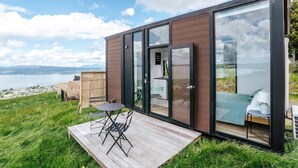 A modern cabin with a deck overlooking a body of water, featuring a glass door entrance and a small outdoor seating area.