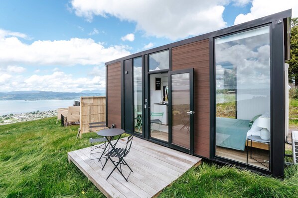 Una cabaña moderna con terraza y vistas a un cuerpo de agua, con puerta de entrada de vidrio y un pequeño espacio de estar al aire libre.