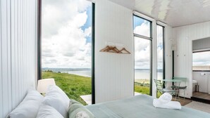 A bedroom with a large window, a bed with white linens, a wooden hanger, and a view of a green landscape.