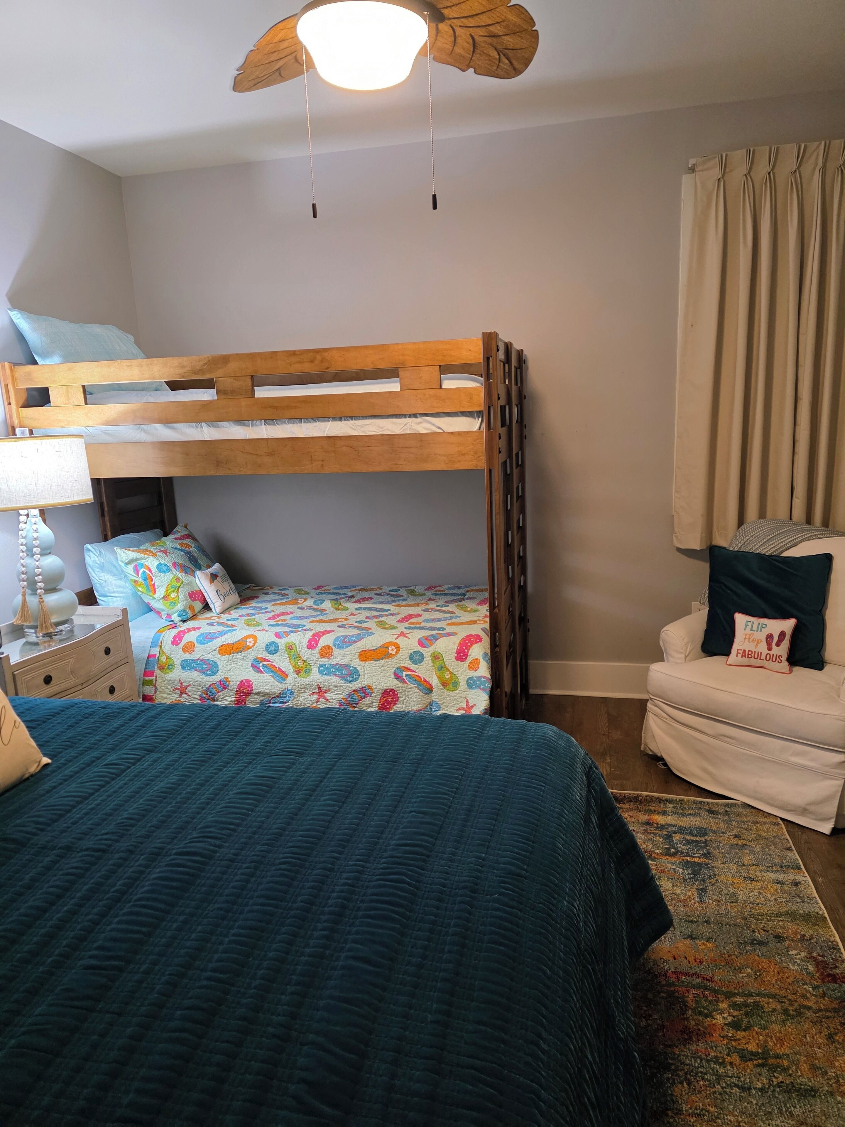 A bedroom with a bunk bed, a bedside table with a lamp, a chair with a cushion, and a ceiling fan.