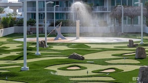 Een aangelegde tuin met een fontein in het midden, omringd door strak gemaaid gras en rotsen, voor een modern gebouw met balkons.