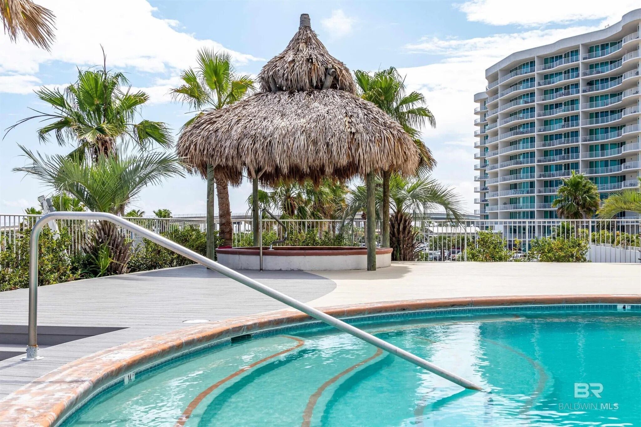 A pool area with a thatched structure, palm trees, and a high-rise building in the background.