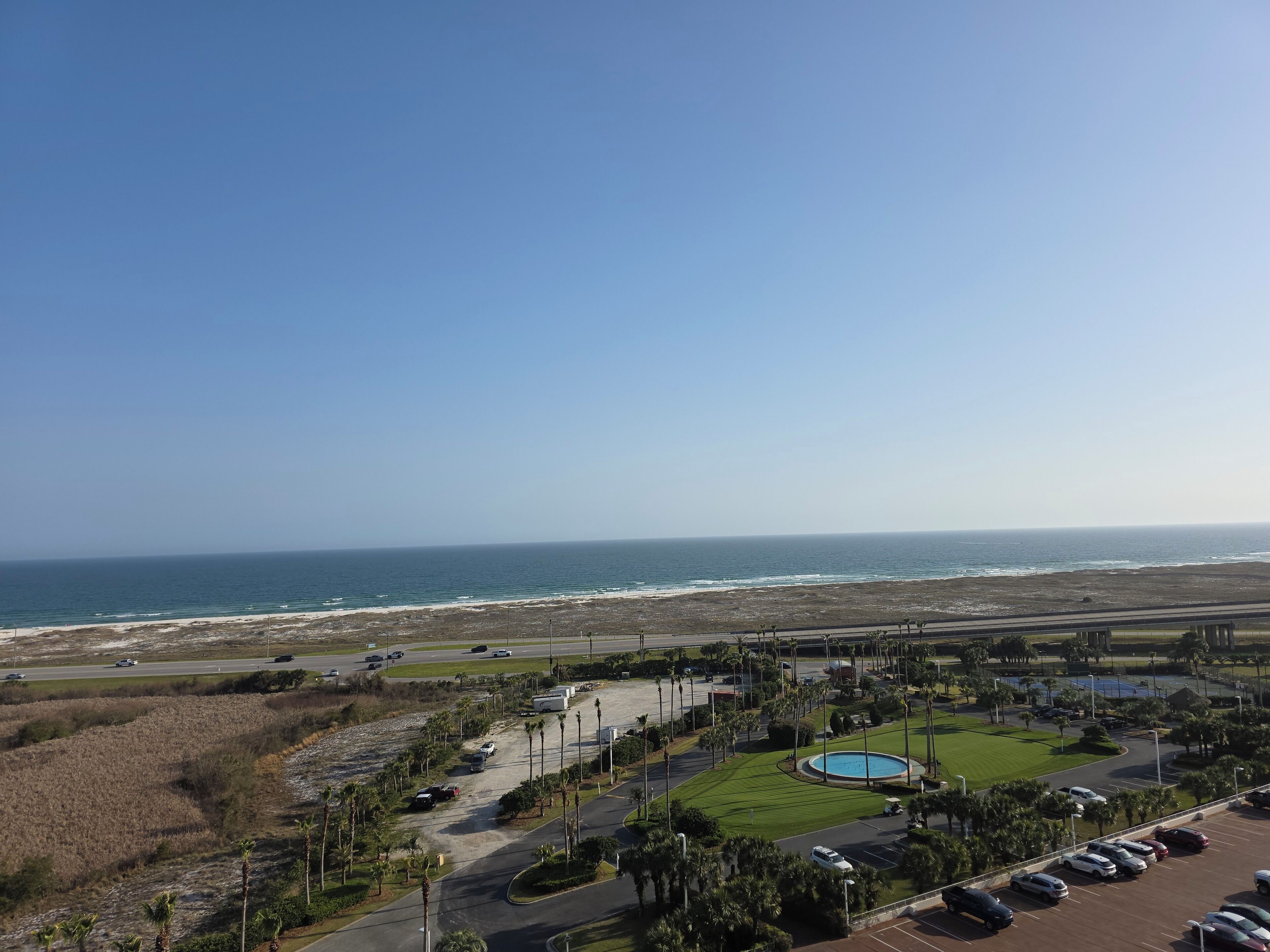 A coastal view with a parking lot, a swimming pool, and a road leading to the beach.