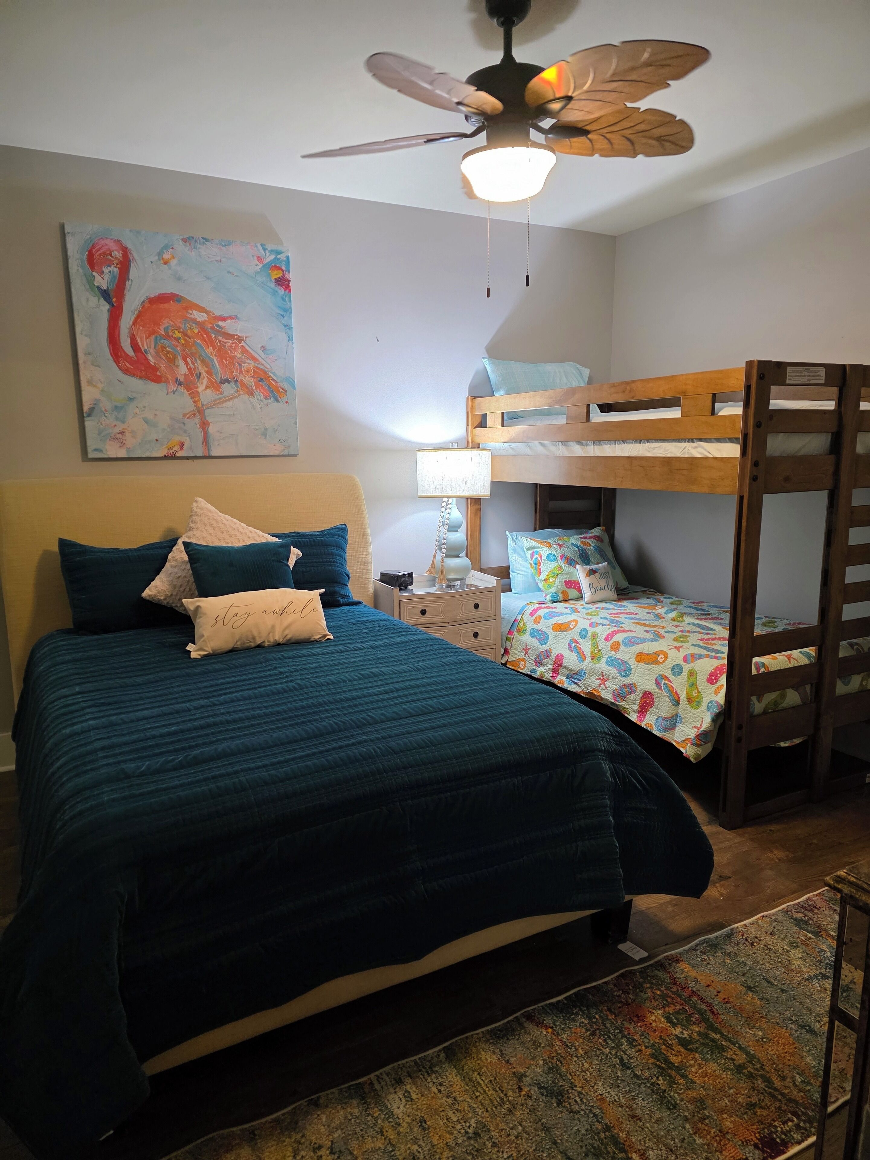 Ein Schlafzimmer mit Etagenbett, einem Bett mit türkisfarbener Bettdecke, Deckenventilator, Lampe und einem Flamingo-Gemälde.