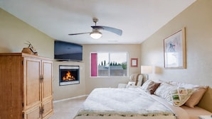 Un dormitorio con cama, chimenea, ventilador de techo, televisor y ventana con vista a árboles.