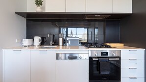 Une cuisine moderne avec des meubles blancs, un lave-vaisselle Bosch, une cuisinière et une grande fenêtre donnant sur un immeuble.
