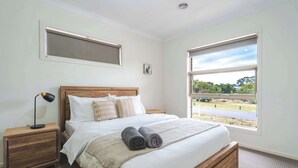 Ein Schlafzimmer mit einem großen Bett, einem Nachttisch, einer Lampe und einem Fenster mit Blick auf eine Straße und Grünflächen.