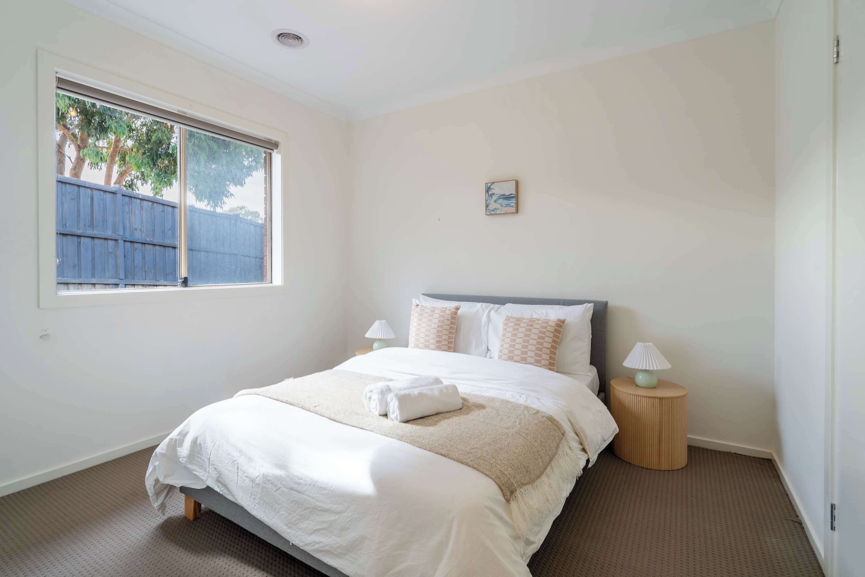 A bedroom with a large bed, two bedside lamps, a window with a view of a fence and trees, and a small painting on the wall.