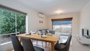 Ein moderner Essbereich mit Holztisch und grauen Stühlen, großen Fenstern mit Blick auf Bäume und einer weißen Kommode mit Fernseher.