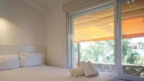 Un dormitorio con cama, almohadas, una ventana y vistas a la vegetación exterior.