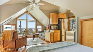 Un dormitorio estilo loft con un ventanal grande, ventilador de techo, kitchenette y vista al océano.
