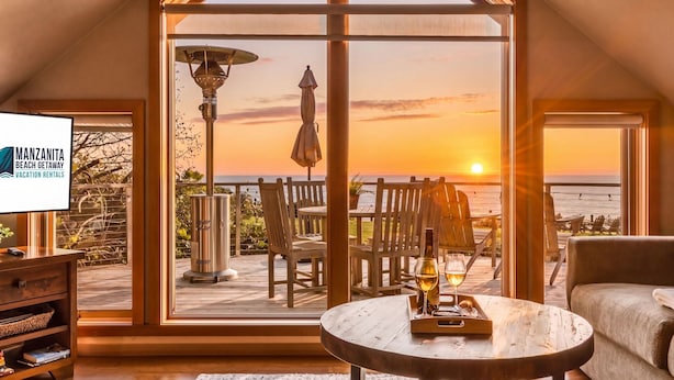 Una habitación con un gran ventanal con vista a un atardecer, una mesa de madera con vasos, y una pantalla de televisión que muestra 'Manzanita Beach Getaway Vacation Rentals'.