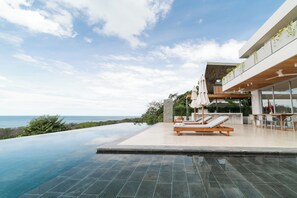 Un área de piscina moderna al aire libre con sillones de descanso y una mesa de comedor, con vistas a un paisaje marino.