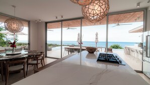 Une cuisine moderne dotée d’un grand îlot central, d’un coin repas offrant une vue sur l’océan et d’une terrasse avec des sièges d’extérieur.