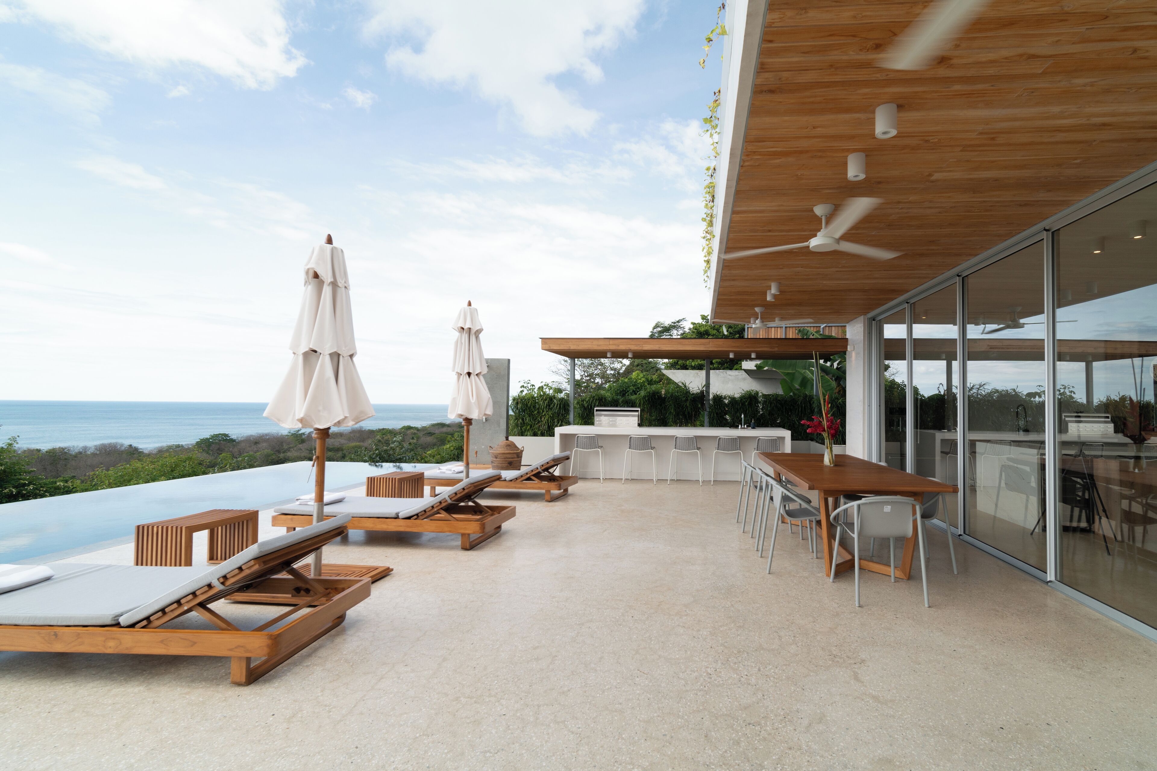 A rooftop terrace with a view of the ocean, wooden lounge chairs, a bar area, and a glass-enclosed space.