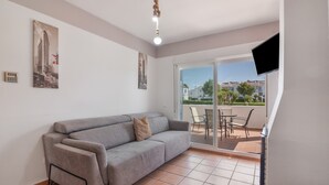 Ein Wohnzimmer mit grauem Sofa, einem an der Wand montierten Flachbildfernseher und einer Schiebetür, die auf eine Terrasse mit Tisch und Stühlen führt.