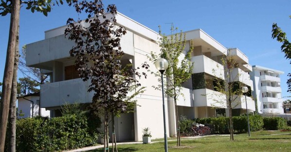 A modern multi-story residential building with balconies, surrounded by greenery and a clear sky.