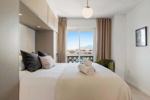 A hotel room with a bed, a chair, a window with curtains, and a view of buildings.