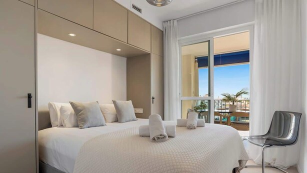 Ett hotellrum med en stor säng, en sittgrupp med en fåtölj och utsikt över en pool och byggnader genom ett stort fönster.