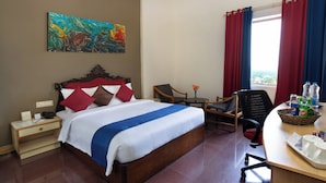 A hotel room with a large bed, a desk with a chair, a small table with a vase, and a window with curtains.