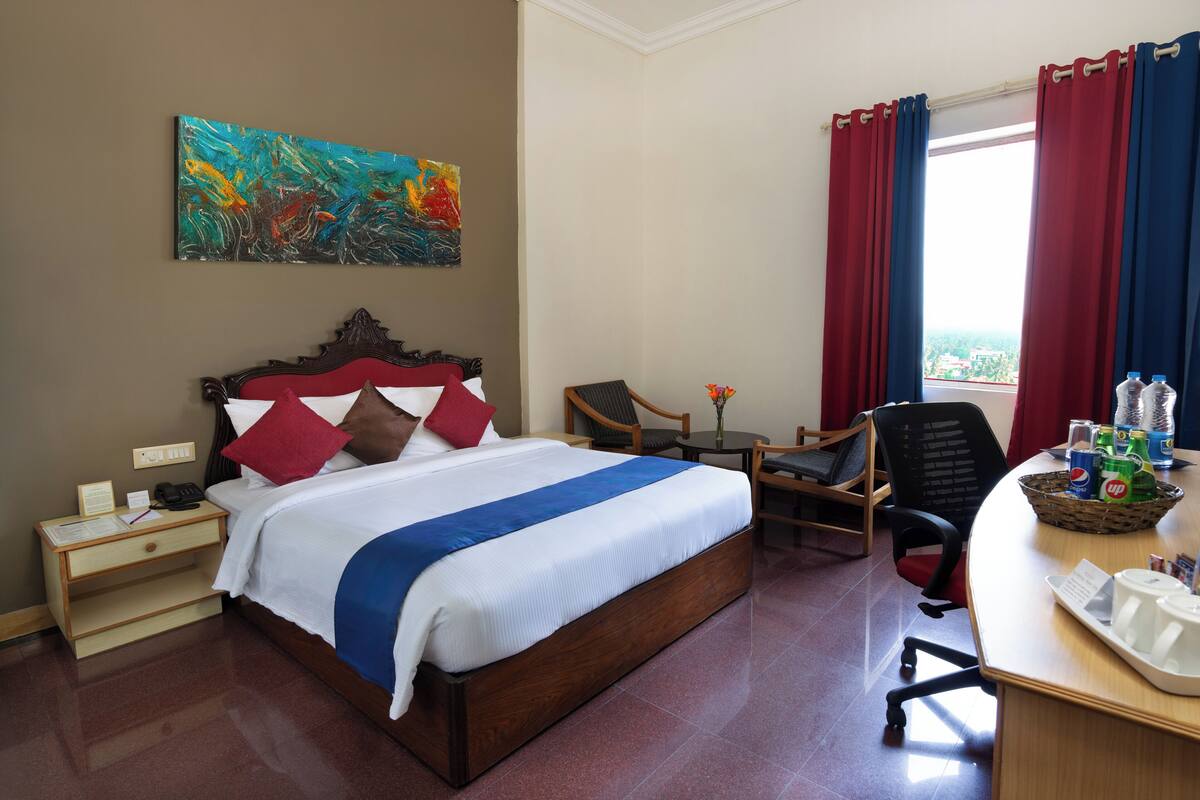 Ein Hotelzimmer mit einem großen Bett, einem Schreibtisch mit Stuhl, einem kleinen Tisch mit Vase und einem Fenster mit Vorhängen.