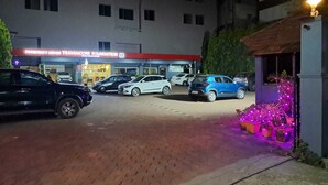 A parking lot with several cars, a building with a sign reading "Presidency Homes Travancore Foundation", and a small structure with purple lights.