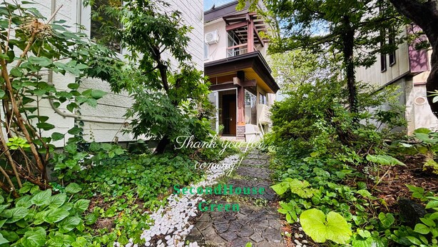 A narrow stone pathway flanked by lush greenery and a building with a wooden balcony.