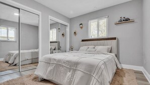 A bedroom with a large bed, a mirror, a window with blinds, and a shelf with a bicycle decoration. - Hot Tub, Fire Pit, Near the Beach! (Edgewater)