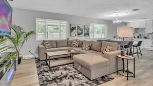 A modern living room with a sectional sofa, a coffee table, and a TV. There is a dining area with a table and chairs. - Hot Tub, Fire Pit, Near the Beach! (Edgewater)