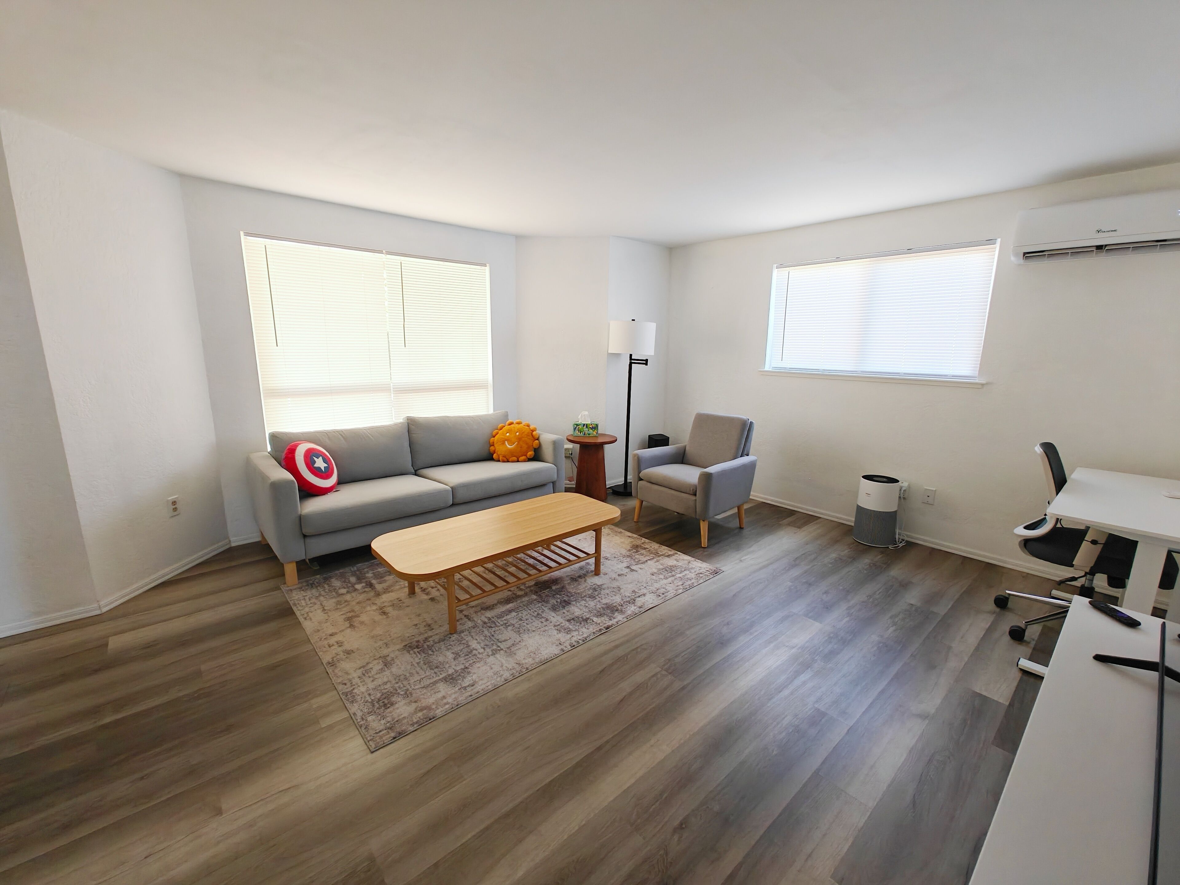 Un salon moderne avec un canapé gris, une table basse en bois, un fauteuil gris et un bureau blanc avec une chaise. Il y a deux fenêtres équipées de stores, une lampe sur pied et une poubelle.