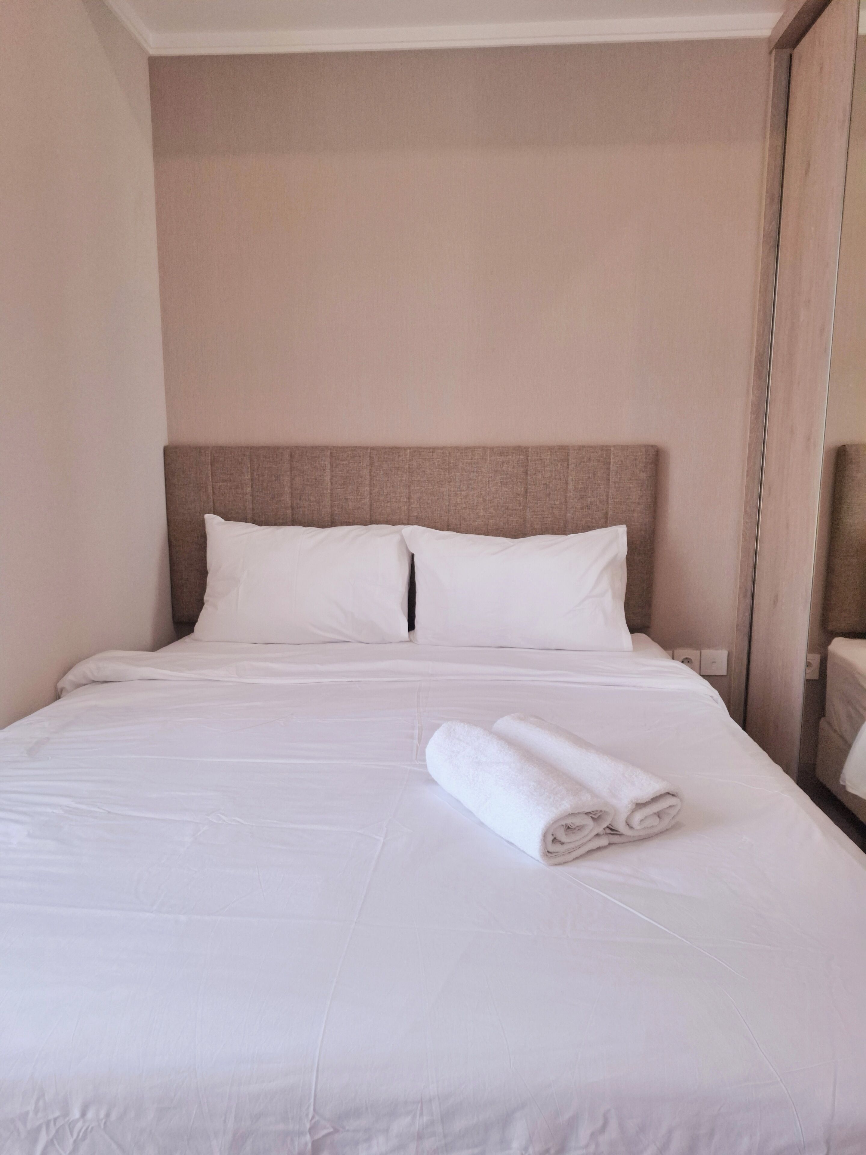 Una cama bien hecha con sábanas y almohadones blancos, una toalla enrollada sobre la cama y un espejo en la pared.