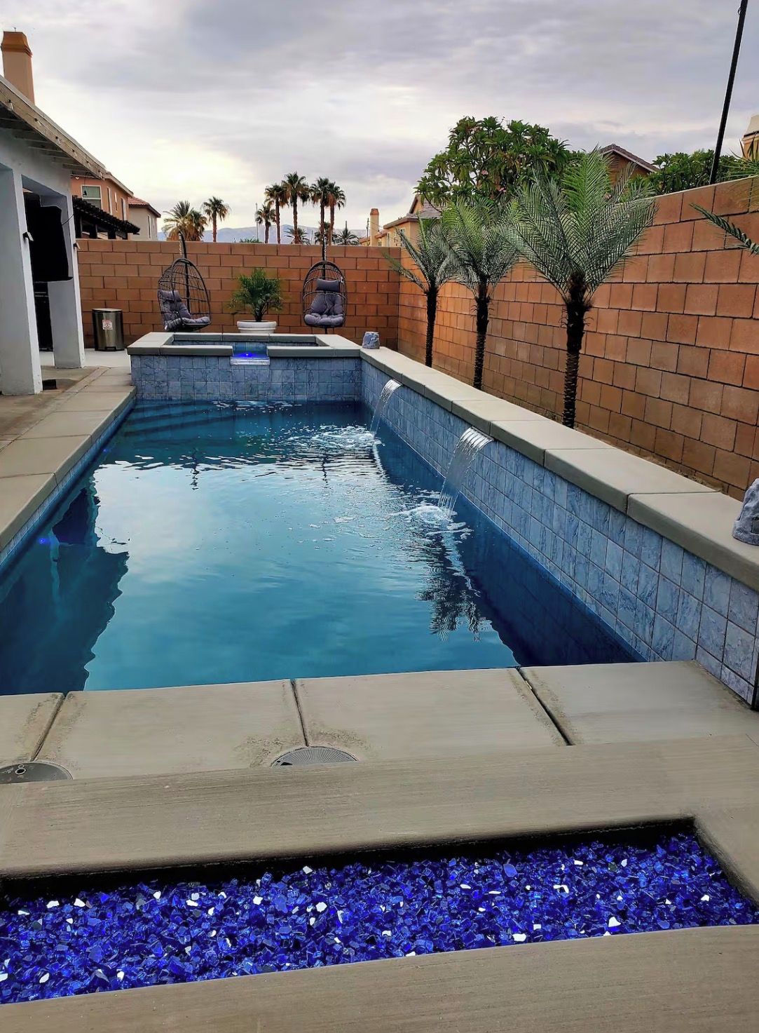 A rectangular swimming pool with a blue tile interior, surrounded by a brick wall and palm trees.