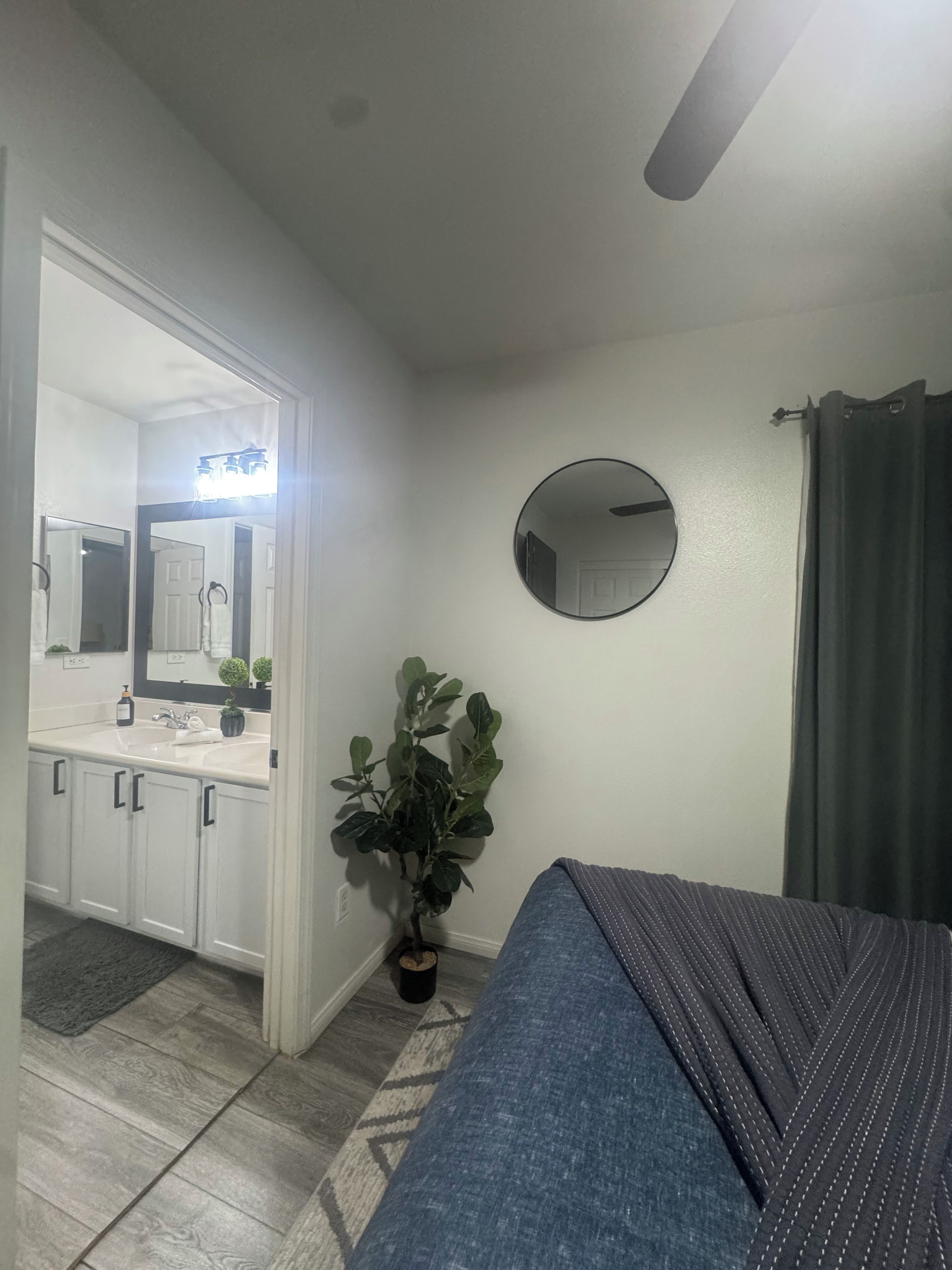 A bedroom with a bed, a round mirror, a potted plant, and a bathroom with a sink and mirror.