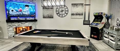 A modern living room with a pool table, a flat-screen TV, a clock, and a pinball machine.